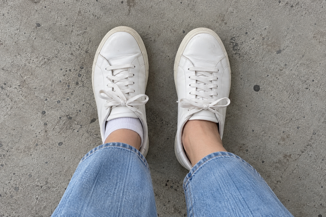 Top down view of white leather sneakers on concrete. The left foot shows a white low cut sock above the collar, the right foot wears a no show sock with nothing visible.
